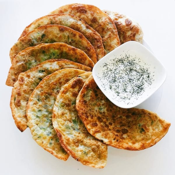Golden-brown Afghan dumplings (Aushak) with green filling, arranged around a white bowl of yogurt sauce.