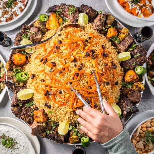 A large platter of Ruz Bukhari's Afghan rice, grilled meats, and vegetables, with a hand serving.